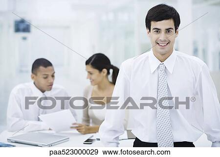 Businessman smiling at camera, colleagues working in background View Large Photo Image Stock Photo - Businessman smiling at camera, colleagues working in background. Fotosearch