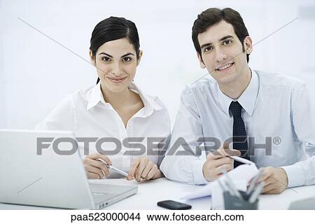 Colleagues sitting together at desk, smiling at camera View Large Photo Image Picture - Colleagues sitting together at desk, smiling at camera. Fotosearch