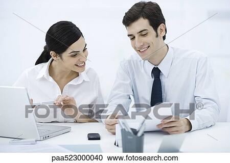 Colleagues sitting together at desk, looking at document, smiling View Large Photo Image Stock Image - Colleagues sitting together at desk, looking at document, smiling. Fotosearch