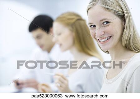 Young professional woman smiling at camera, colleagues working in background View Large Photo Image Stock Image - Young professional woman smiling at camera, colleagues working in background. Fotosearch