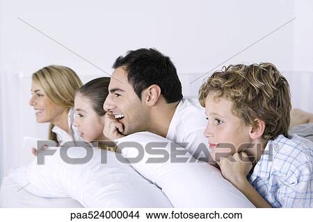 Picture - Family lying on bed, watching television, side view. Fotosearch