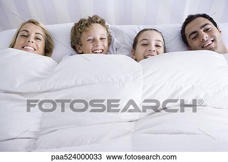 Stock Image - Family lying together in bed beneath comforter, smiling at camera. Fotosearch