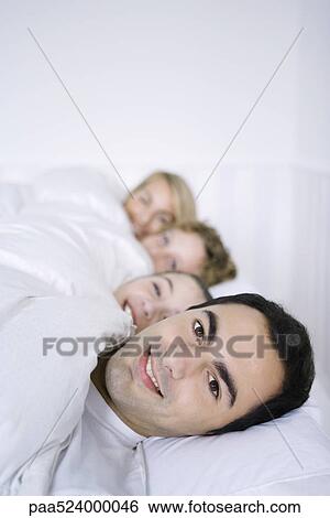 Stock Photograph - Man lying in bed with his family, smiling at camera. Fotosearch