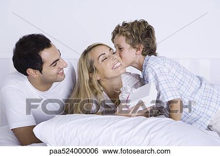 Woman lying in bed with husband and son, holding gift, boy kissing her cheek View Large Photo Image Stock Photograph - Woman lying in bed with husband and son, holding gift, boy kissing her cheek. Fotosearch