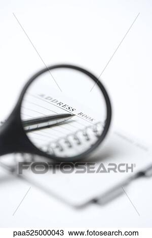 Stock Image - Magnifying glass over address book and pen, close-up. Fotosearch