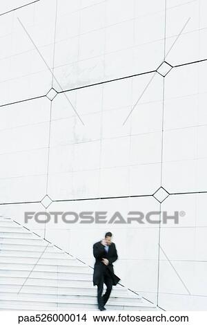 Picture - Businessman descending outdoor stairs, talking on cell phone, holding coat closed against the wind. Fotosearch