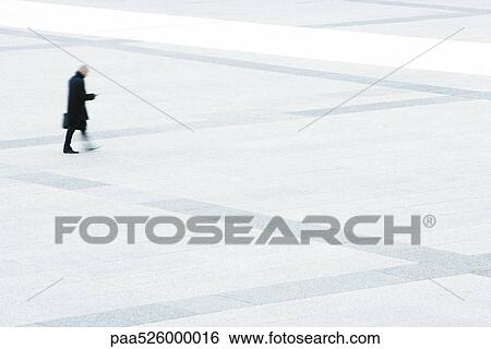 Businessman looking at his cell phone while walking across public square View Large Photo Image Stock Photograph - Businessman looking at his cell phone while walking across public square. Fotosearch