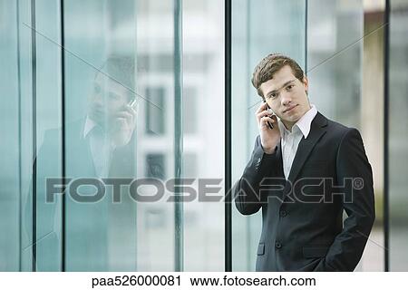 Businessman standing by glass wall talking on cell phone View Large Photo Image Stock Image - Businessman standing by glass wall talking on cell phone. Fotosearch