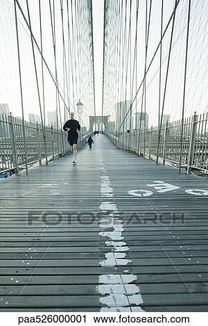Stock Image - Jogger running across pedestrian walkway of Brooklyn Bridge in New York City, Manhattan skyline in distance. Fotosearch