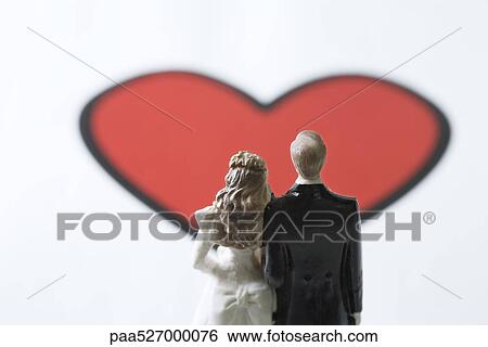 Stock Photograph - Miniature bride and groom standing in front of large heart graphic. Fotosearch