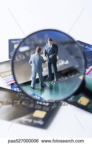 Stock Photograph - Miniature businessmen shaking hands on pile of credit cards, viewed through magnifying glass. Fotosearch