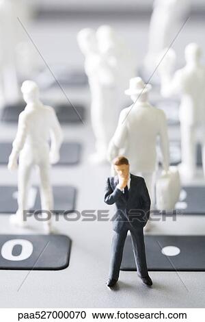 Stock Image - Miniature man figure in suit standing with hand under chin, white figures in the background. Fotosearch