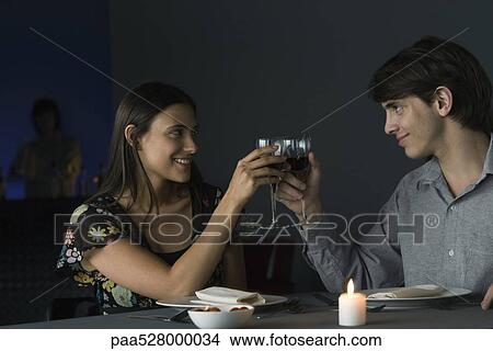Picture - Couple clinking glasses at restaurant, smiling at each other. Fotosearch