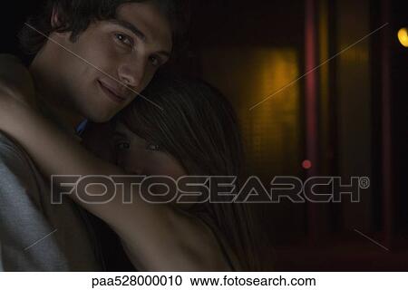 Couple embracing in darkly lit room, looking over shoulder at camera View Large Photo Image Stock Image - Couple embracing in darkly lit room, looking over shoulder at camera. Fotosearch