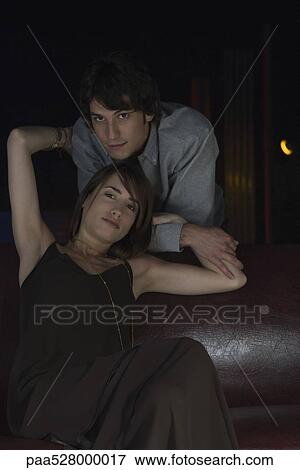 Stock Photo - Couple smiling at camera, woman sitting with arms raised embracing man standing behind. Fotosearch