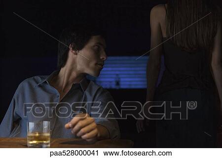 Stock Image - Glass of whiskey on table, man sitting and looking away, rear view of woman standing in the dark. Fotosearch