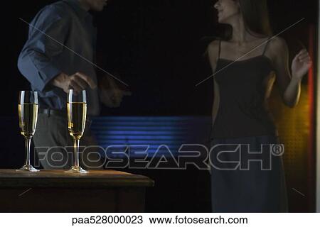 Stock Image - Glasses of champagne, couple dancing in darkly lit background. Fotosearch
