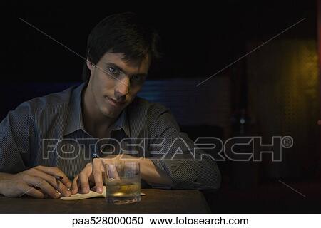 Stock Image - Man sitting in bar with pen, notebook and glass of whiskey, looking at camera. Fotosearch