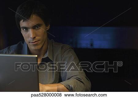 Man sitting in darkly lit room at table with laptop, looking at camera, portrait View Large Photo Image Stock Photo - Man sitting in darkly lit room at table with laptop, looking at camera, portrait. Fotosearch