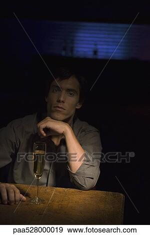 Stock Photo - Man sitting in the dark, hand under chin, spotlight on glass of champagne on table . Fotosearch