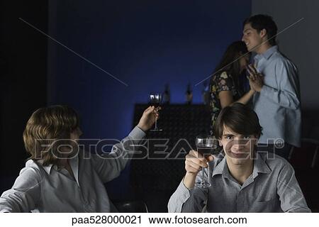 Stock Image - Men raising glasses, one looking over shoulder at dancing couple, other smiling at camera. Fotosearch