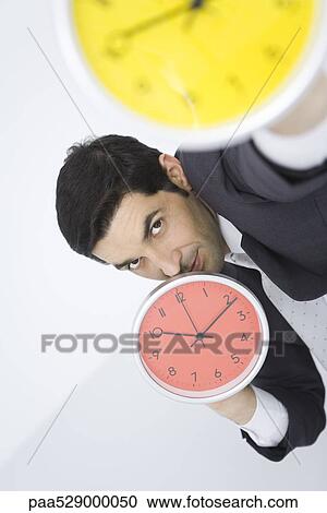Businessman holding up clocks, looking at camera View Large Photo Image Stock Image - Businessman holding up clocks, looking at camera. Fotosearch