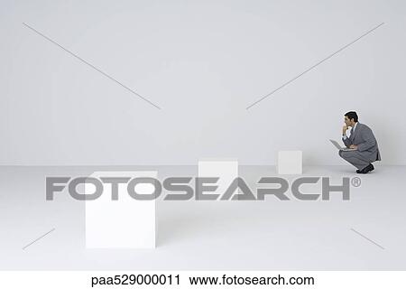 Stock Image - Businessman squatting using laptop, looking at empty pedestals. Fotosearch