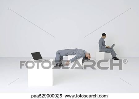 Identical businessmen, one using laptop while the other lies on back, exhausted View Large Photo Image Stock Photography - Identical businessmen, one using laptop while the other lies on back, exhausted. Fotosearch