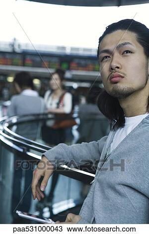 Stock Image - Young man leaning against railing, holding cell phone. Fotosearch