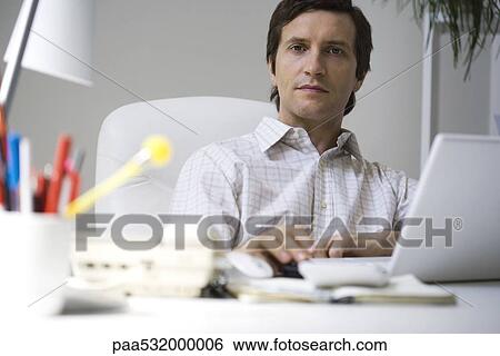 Stock Photograph - Businessman sitting at desk, working on laptop. Fotosearch