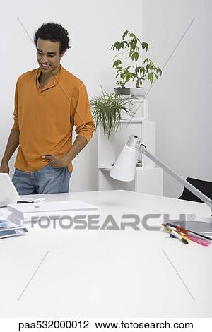 Casual man standing by desk with hand in pocket, looking down at laptop View Large Photo Image Stock Image - Casual man standing by desk with hand in pocket, looking down at laptop. Fotosearch