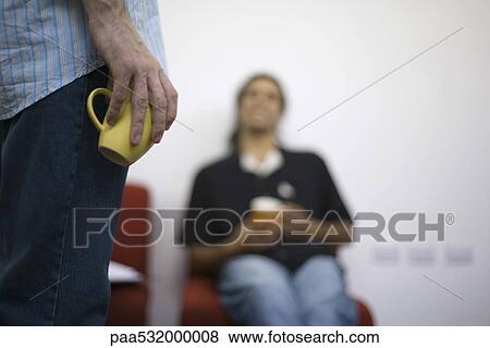 Man casually holding coffee cup at side, colleague sitting in background View Large Photo Image Stock Photo - Man casually holding coffee cup at side, colleague sitting in background. Fotosearch