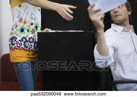 Stock Photo - Man handing document to colleague standing by filing cabinet. Fotosearch