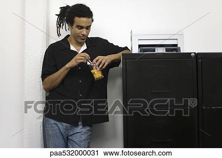 Stock Image - Man leaning against filing cabinet, stirring drink. Fotosearch