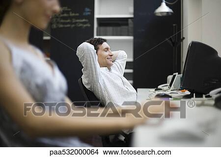 Picture - Man relaxing at desk, woman working in foreground. Fotosearch