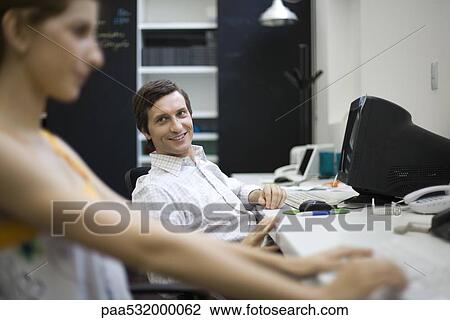 Stock Image - Man sitting at desk, smiling at female colleague. Fotosearch