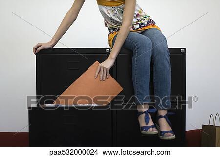 Picture - Woman sitting on filling cabinets, removing file from drawer, cropped. Fotosearch