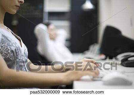 Stock Photo - Woman typing on keyboard, colleague relaxing in background. Fotosearch