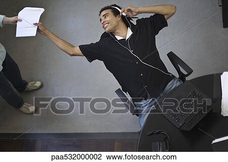 Stock Image - Young office worker leaning back in chair to receive document from colleague, high angle view. Fotosearch