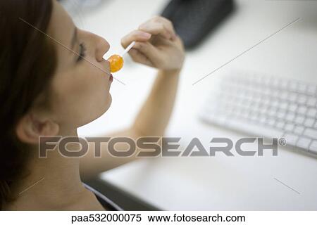 Stock Photography - Young woman sitting at desk, eating lollipop as she works. Fotosearch