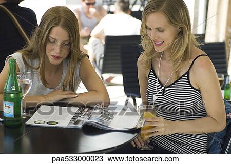 Stock Image - Two young female friends sitting at outdoor cafe, looking at magazine together. Fotosearch