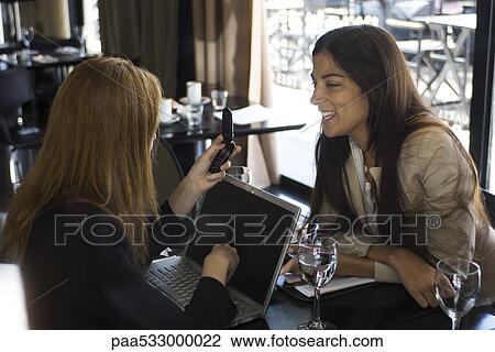 Stock Image - Two young women sitting in cafe, chatting, one using laptop and cell phone. Fotosearch