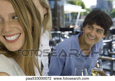 Stock Image - Young friends sitting at sidewalk cafe, looking over shoulders and smiling. Fotosearch
