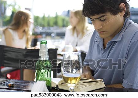 Stock Image - Young man sitting at outdoor cafe, reading book and having a beer. Fotosearch