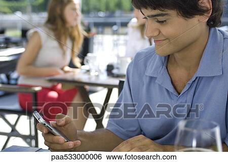Stock Photograph - Young man sitting at outdoor cafe, using cell phone. Fotosearch