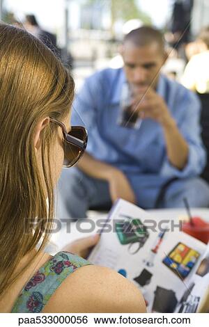 Stock Photograph - Young woman sitting at a sidewalk cafe, reading catalogue, over the shoulder view. Fotosearch
