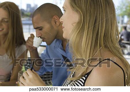 Stock Image - Young woman sitting with friends at sidewalk cafe, close-up. Fotosearch