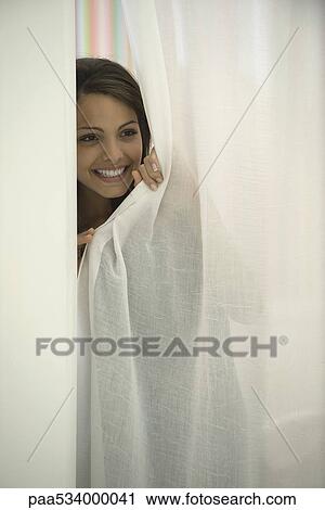 Stock Image - Woman peeking out curtain, smiling. Fotosearch