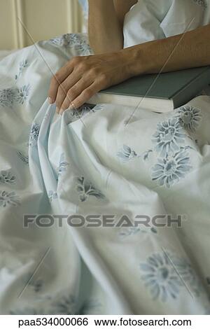 Stock Photograph - Woman sitting in bed with hand resting on book, cropped view. Fotosearch