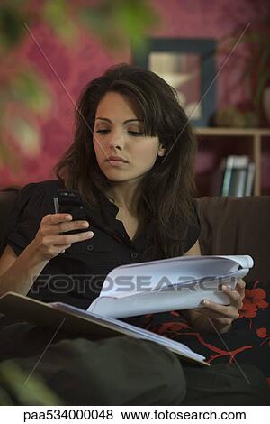 Stock Photo - Woman sitting on sofa with document, looking down at cell phone. Fotosearch